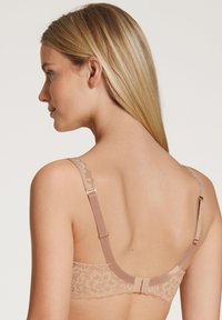 Reggiseno con schiena in pizzo color nude, con bordi di pizzo a scallop, spalline regolabili e dettagli in hardware dorato sulla chiusura. Tessuto complessivamente liscio.