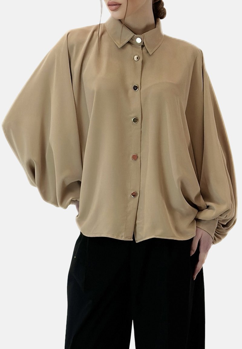 Blouse beige oversize avec un col classique, quatre grands boutons, des manches volumineuses et une texture lisse, associée à un pantalon noir.