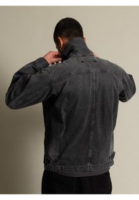 Man som justerar kragen på en urblekt svart denimjacka, vänd bort från en enfärgad beige bakgrund.