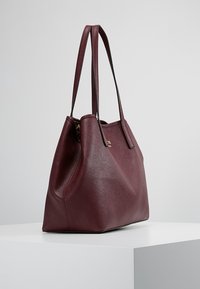 Dieprode leren draagtassen met dubbele schouderbanden rustend op een wit voetstuk tegen een effen achtergrond.