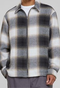 Mężczyzna ubrany w czarną, białą i beżową kurtkę z zamkiem na kratę, pod którą ma białą koszulę oraz szare spodnie, z rękami w kieszeniach kurtki.