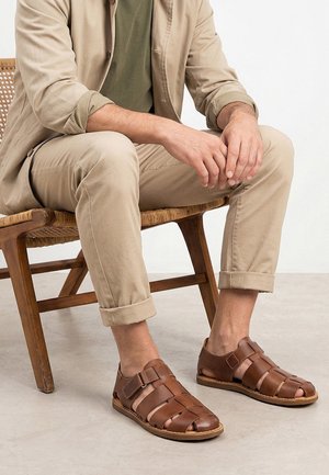 Mężczyzna siedzący na drewnianym krześle, ubrany w beżowe spodnie, jasnobrązową kurtkę oraz brązowe, skórzane sandały zamknięte na palcach z rzepami.