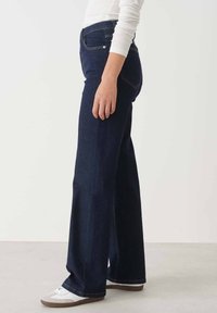 Jeans a gamba larga di un blu scuro, realizzati in denim, presentano dettagli di cucitura discreti e tasche posteriori. Abbinati a scarpe bianche.