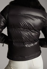 Zwarte pufferjack met een glanzende, gevoerde textuur, een hoge kraag met een bontaccent en een getailleerde taille. Ritssluiting aan de zoom.