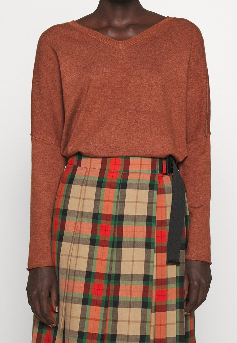 Personne portant un pull à col V couleur rouille rentré dans une jupe plissée à carreaux taille haute avec des tons rouges, verts, beiges et orange.