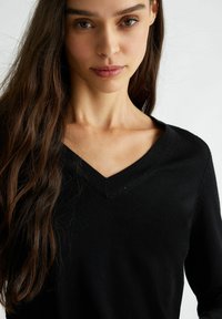 Maglione nero a scollo a V con lavorazione a maglia testurizzata, maniche lunghe e dettagli ad apertura sul colletto. Tessuto liscio con una leggera lucentezza.