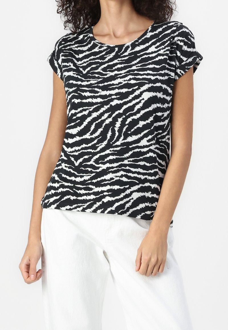 Femme portant un haut à manches courtes rayé zèbre noir et blanc, associé à un pantalon blanc ample, debout devant un fond uni.