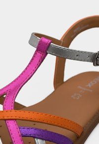 Detailné zábery remienkov sandál v metalickej ružovej, striebornej, oranžovej a fialovej farbe na hnedom podrážke s prackou na zapínanie.