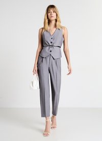 Graue maßgeschneiderte Weste mit V-Ausschnitt und drei Knöpfen, kombiniert mit schmalen Hosen. Sie verfügt über einen schwarzen Gürtel, eine weiße Handtasche und sandalen mit Riemchen.