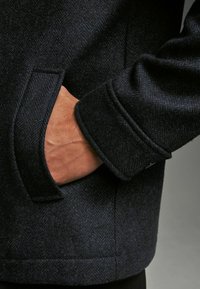 Marineblauer Fischgrät-Wollmantel mit einer Seitentasche und Detail an den Bündchen. Der Stoff ist strukturiert und die Tasche ist so positioniert, dass die Hand hineingelegt werden kann.