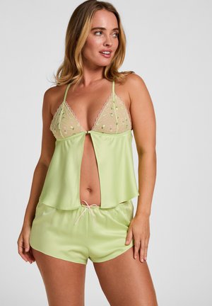 Vrouw die een lichtgroene satijnen slaapset draagt met geborduurde doorschijnende cups en bijpassende korte broek, vastgebonden met een roze strik, poseert tegen een effen achtergrond.