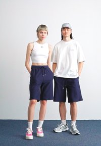 Top court blanc coupé, shorts rayés bleu marine, baskets roses sur un modèle ; t-shirt blanc oversize et shorts bleu marine sur l'autre, portant une casquette grise.