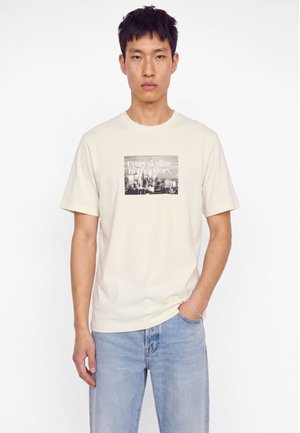 Homme portant un t-shirt crème avec un graphisme de silhouette de ville noire et le texte "Every skyline has a story", assorti à un jean bleu clair et un bracelet argenté.