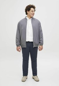 Grijze bomberjack met geribde manchetten, witte overhemd met kraag, marineblauwe broek en beige instappers, staand op een effen achtergrond.