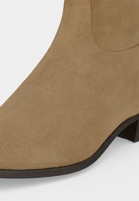 Gros plan d'une botte cheville en daim marron clair avec un talon noir bas et un bout arrondi sur fond blanc.