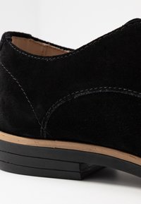 Chaussure en daim noir avec des accents cousus et un petit talon. La semelle présente un design superposé avec un détail en tan sur le bord.