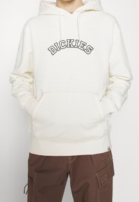 Sudadera blanca con "DICKIES" bordado en negro en el pecho. Cuenta con un bolsillo frontal y capucha con cordones, hecha de un tejido suave.