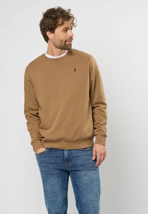 Beige sweatshirt met ronde halslijn en ribgebreide manchetten, voorzien van een klein marineblauw logo op de borst, gecombineerd met blauwe jeans.