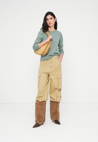 Πράσινο πουλόβερ, μπεζ παντελόνι cargo με τσέπες, καφέ χνουδωτές καλύψεις ποδιών και καφέ μυτερά παπούτσια· κίτρινη τσάντα με λεπτομέρειες φουντών.