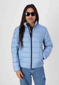Ljusblå uppblåsbar jacka med dragkedja fram, med horisontell sömnad och en logotyp på dragkedjan. Matchas med mörka jeans och solglasögon.