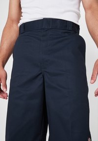 Homme portant un pantalon de travail bleu marine et un débardeur blanc, debout les mains détendues le long du corps.