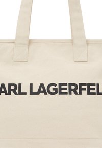 Sac cabas en toile beige clair avec un texte noir audacieux : "KARL LAGERFELD." Poignées solides cousues, forme rectangulaire, et base plate.