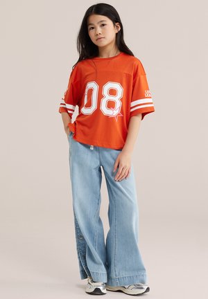 Ragazza giovane in piedi che indossa una maglietta sportiva arancione oversize con il numero "08", jeans larghi azzurri chiari e sneakers bianche su uno sfondo semplice.
