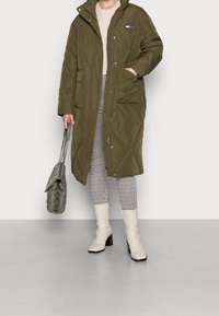 Abrigo largo acolchado verde oliva con cuello alto, botones a presión y bolsillos laterales. Combinado con leggings de pata de gallo y botas blancas de tacón.