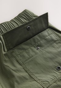 Pantaloncini in tessuto verde con motivo strutturato, vita elastica e chiusura con bottoni a pressione. Presentano una tasca posteriore con ferramenta nera.