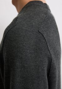 Nahaufnahme einer dunkelgrauen gestrickten Pullover-Schulter und Oberarm einer Person mit heller Haut vor einem schlichten hellen Hintergrund.