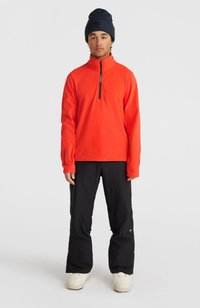 Oranje fleece pullover met halverwege ritssluiting, lange mouwen en zwarte skibroek. Model draagt een donkere beanie; schoeisel bestaat uit chunky witte sneakers.