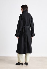 Long manteau en denim noir avec une ceinture, des manches larges et un grand col. Porté par-dessus un pantalon vert clair et des bottes noires à talons, vu de dos.