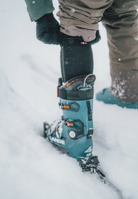Scarponi da sci di un verde scuro con accessori arancioni, caratterizzati da un design robusto, fibbie regolabili e una suola testurizzata, ambientati in un contesto innevato.