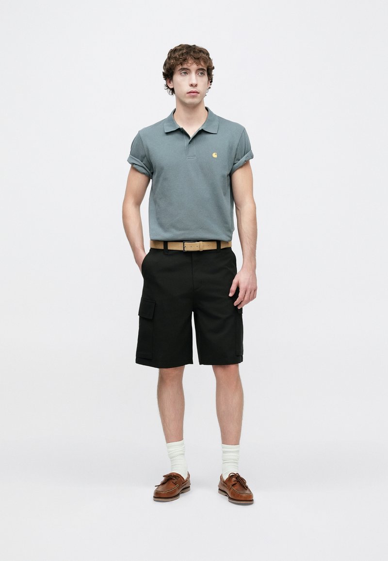 Polo gris à manches retroussées avec un logo jaune, associé à un short cargo noir. Ceinture marron, chaussettes claires et chaussures bateau marron.