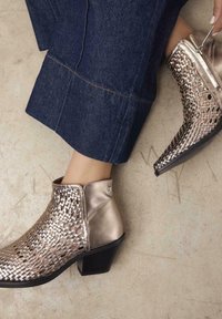 Botines dorados metálicos con detalles trenzados, textura suave, tacón cuadrado pequeño y cremallera lateral, combinados con jeans de pierna ancha.