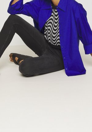 Osoba siedząca ze skrzyżowanymi nogami, ubrana w niebieską koszulę zapinaną na guziki, czarno-biały wzorzysty top, czarne spodnie i czarne sandały.