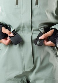 Tactical jumpsuit in soft green fabric, featuring adjustable black straps and hardware, with textured, reinforced areas visible.