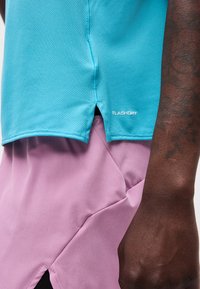 Prim-plan cu o persoană purtând un top turcoaz texturat cu textul „FLASHDRY” și pantaloni scurți liliachii deschis, arătând o parte a brațului tatuat cu pielea închisă la culoare.