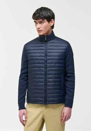 Man draagt een marineblauwe gewatteerde jas met rits en lange mouwen, met beige broek, kijkt opzij tegen een effen achtergrond.