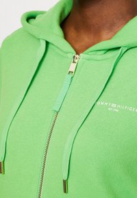 Sudadera verde con cremallera hecha de una tela suave, con cremallera dorada y cordones, con el logo "Tommy Hilfiger" bordado en el frente.