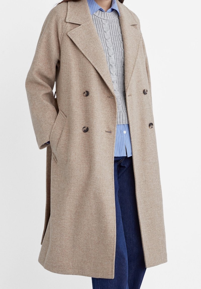 Beżowy wełniany płaszcz dwurzędowy z klapami, przednimi kieszeniami i ciemnymi guzikami. Noszony na szarym swetrze w warkocze i niebieskiej koszuli w paski.