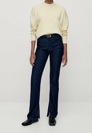 Personne portant une chemise crème à manches longues, un jean foncé taille haute avec des fentes à l'avant, une ceinture noire et des chaussures noires à bout carré, debout contre un mur blanc.