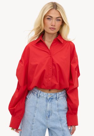 Femme aux longs cheveux blonds portant une chemise rouge ample à boutons et un jean bleu clair taille haute, debout devant un fond blanc uni.