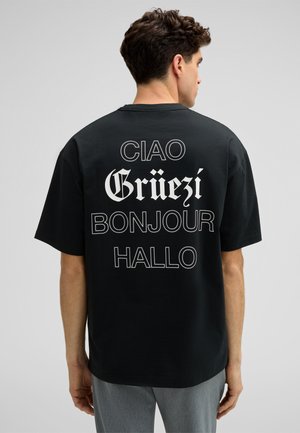 Schwarzes T-Shirt mit kurzen Ärmeln, das auf der Rückseite mehrfarbigen Text zeigt: "Ciao", "Grüezi", "Bonjour" und "Hallo" in verschiedenen Schriftarten.