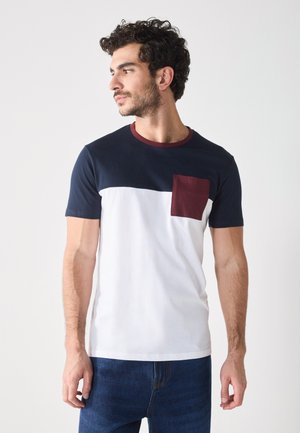 Camiseta de manga corta con una sección superior de color azul marino, una sección inferior blanca y un bolsillo burdeos. Hecha de algodón y con un escote redondo.