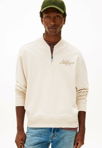 Felpa beige con collo con zip, polsini e orlo a coste, con logo ricamato sul petto, indossata sopra una maglia a righe.