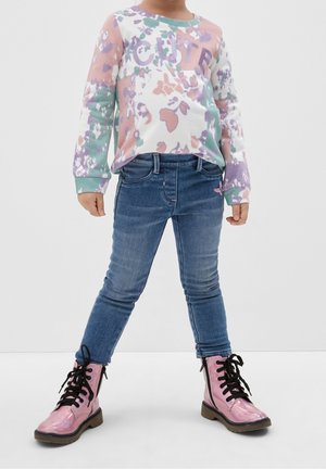 Sweatshirt à manches longues multicolore avec des motifs floraux, skinny jeans en denim bleu, et bottines à lacets roses brillantes.