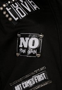 Tessuto nero decorato con diversi patch con testi, tra cui "NO PAIN GAIN" e "L'ARTE PRIMA DI TUTTO", arricchito da borchie e paillettes.
