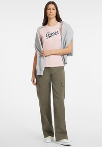 Jasnoróżowa koszulka z krótkim rękawem z granatowym logo "Guess", zestawiona z oliwkowymi spodniami cargo i szarym kardiganem narzuconym na ramionach.