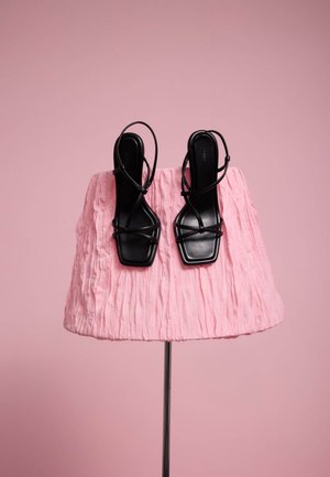 Zwarte sandalen met smalle bandjes en een vierkante neus, tentoongesteld op een roze gestructureerde stof op een metalen standaard tegen een roze achtergrond.